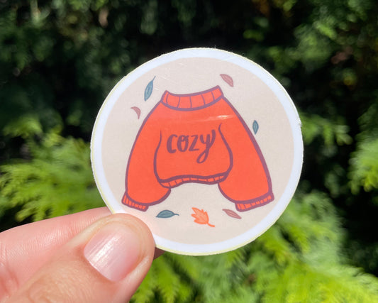 Circular sticker with a red sweater design held by a hand against a green leafy background