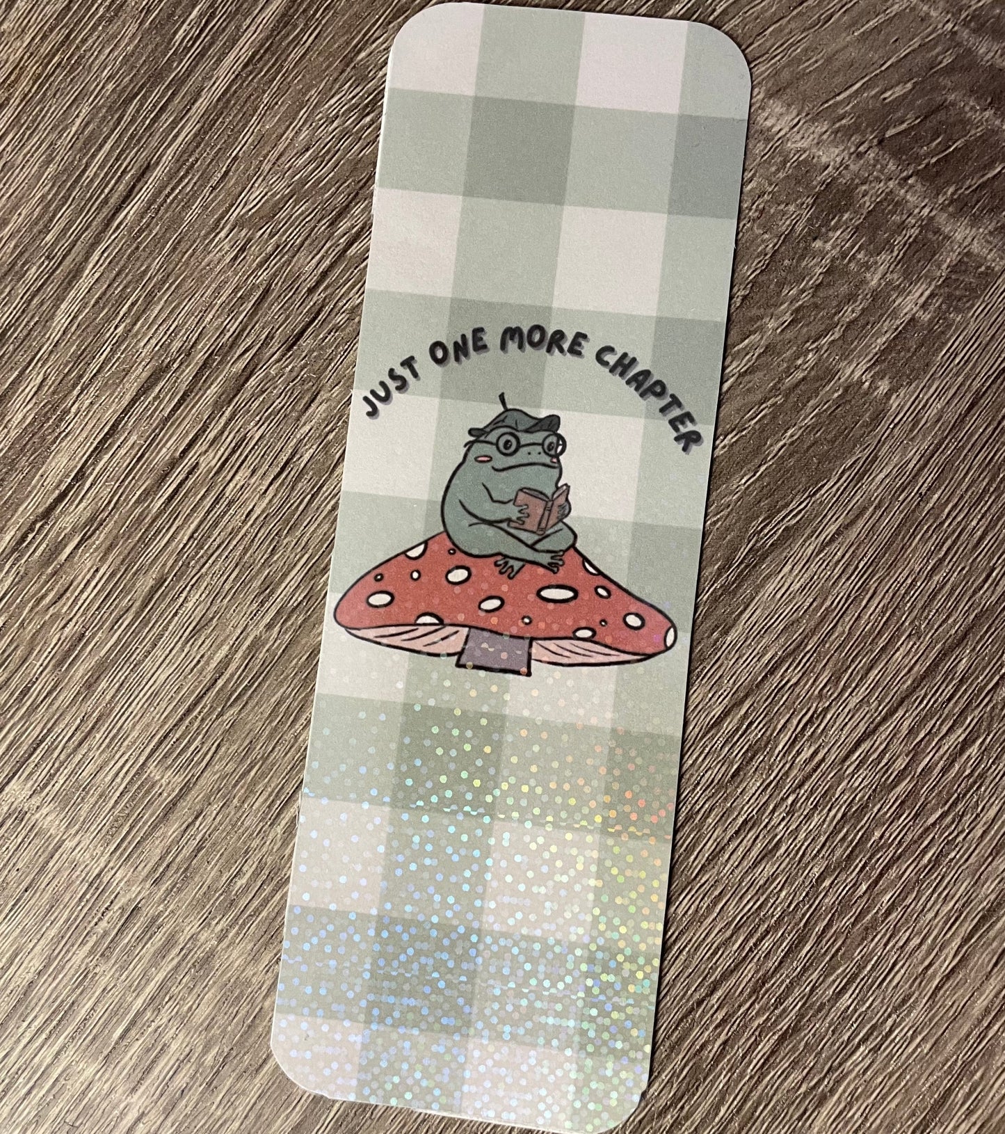 Bookmark with a cartoon character on a mushroom and text 'Just one more chapter' on a checkered background.