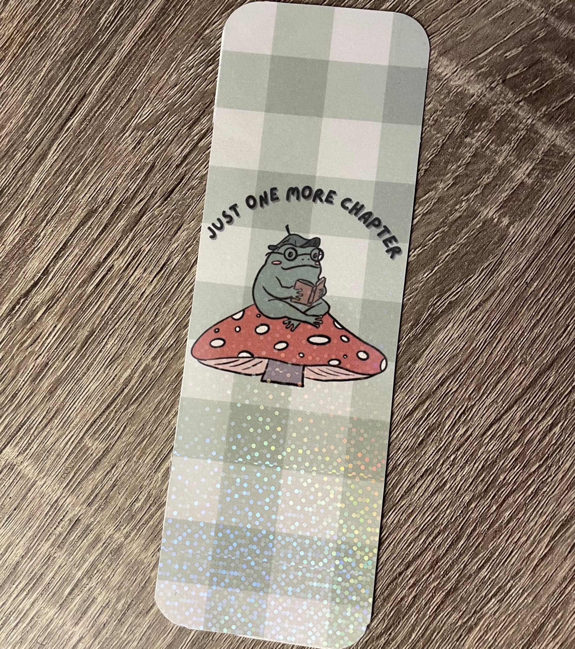 Bookmark with a cartoon character on a mushroom and text 'Just one more chapter' on a checkered background.