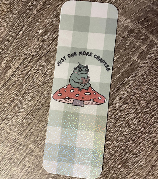 Bookmark with a cartoon character on a mushroom and text 'Just one more chapter' on a checkered background.