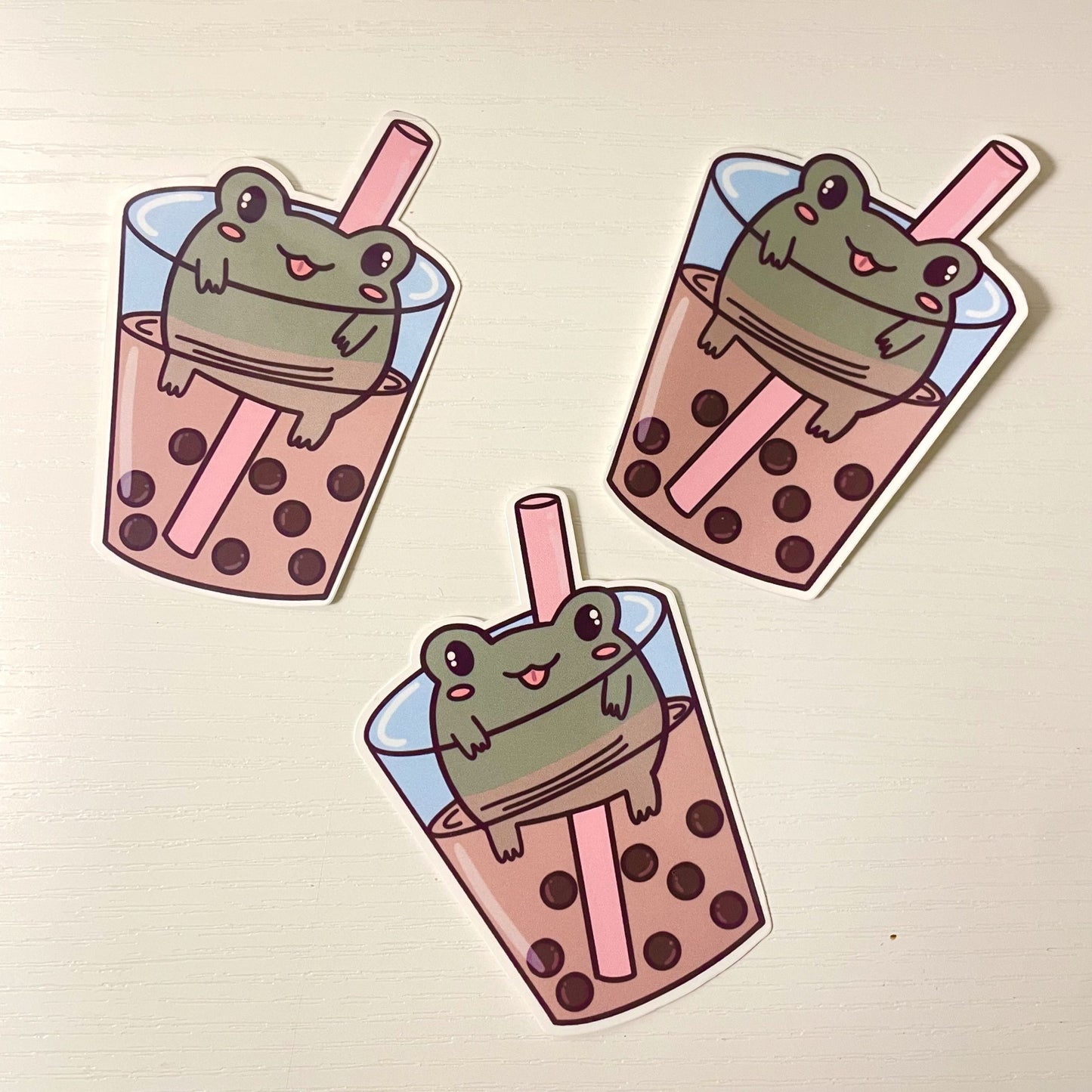 Cute green cartoon frog floating inside a cup of boba milk tea with tapioca pearls and a pink straw.
