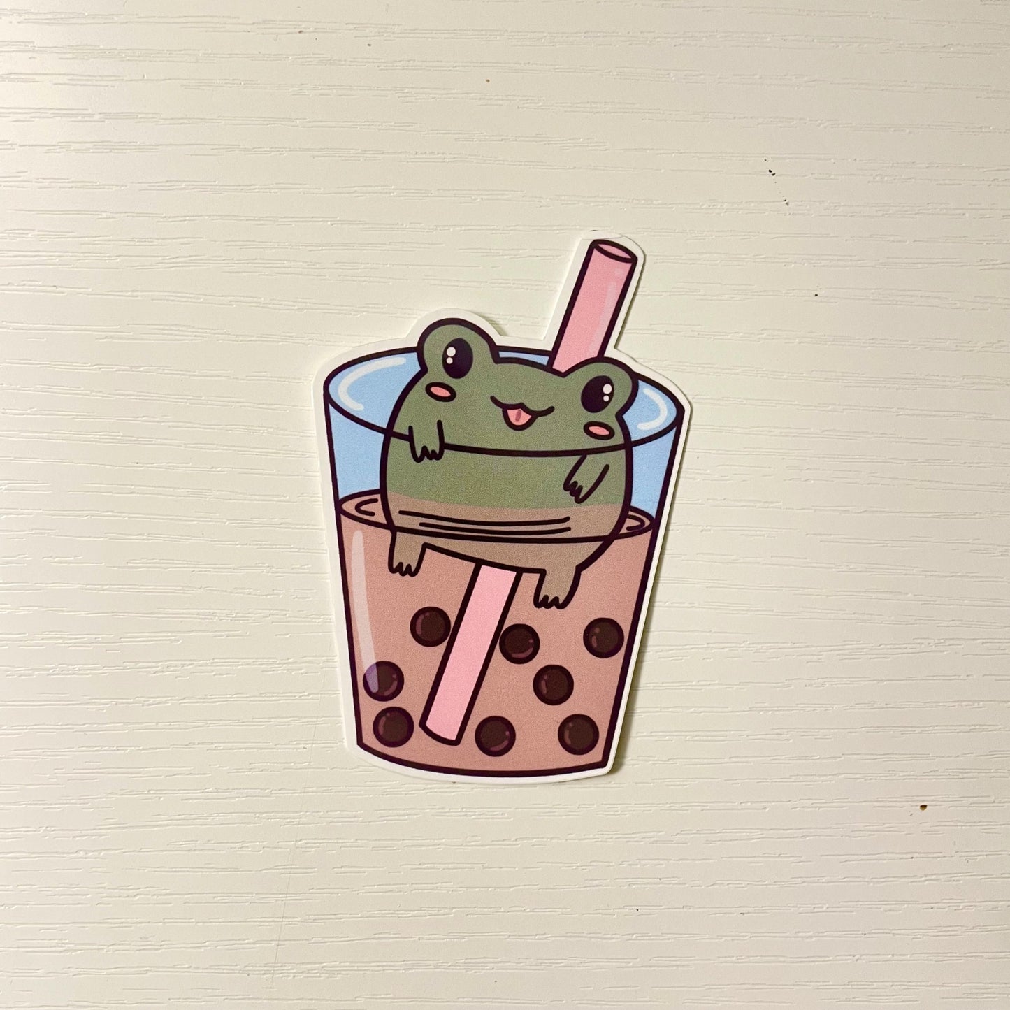Cute green cartoon frog floating inside a cup of boba milk tea with tapioca pearls and a pink straw.