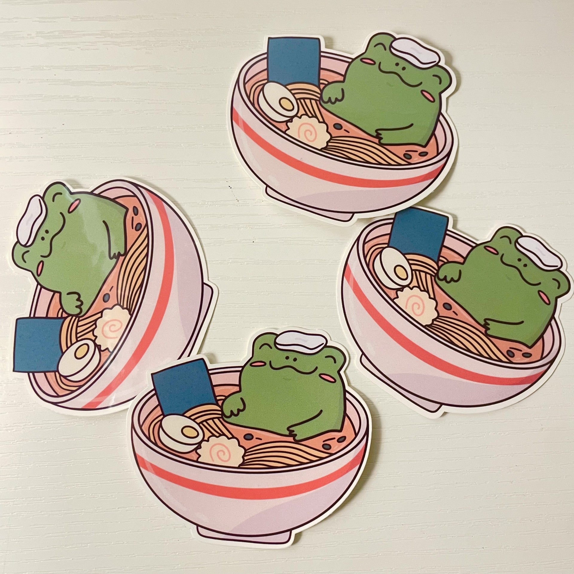 Cute green cartoon frog relaxing in a bowl of ramen noodles with a white towel on its head, a soft boiled egg, and fish cake.