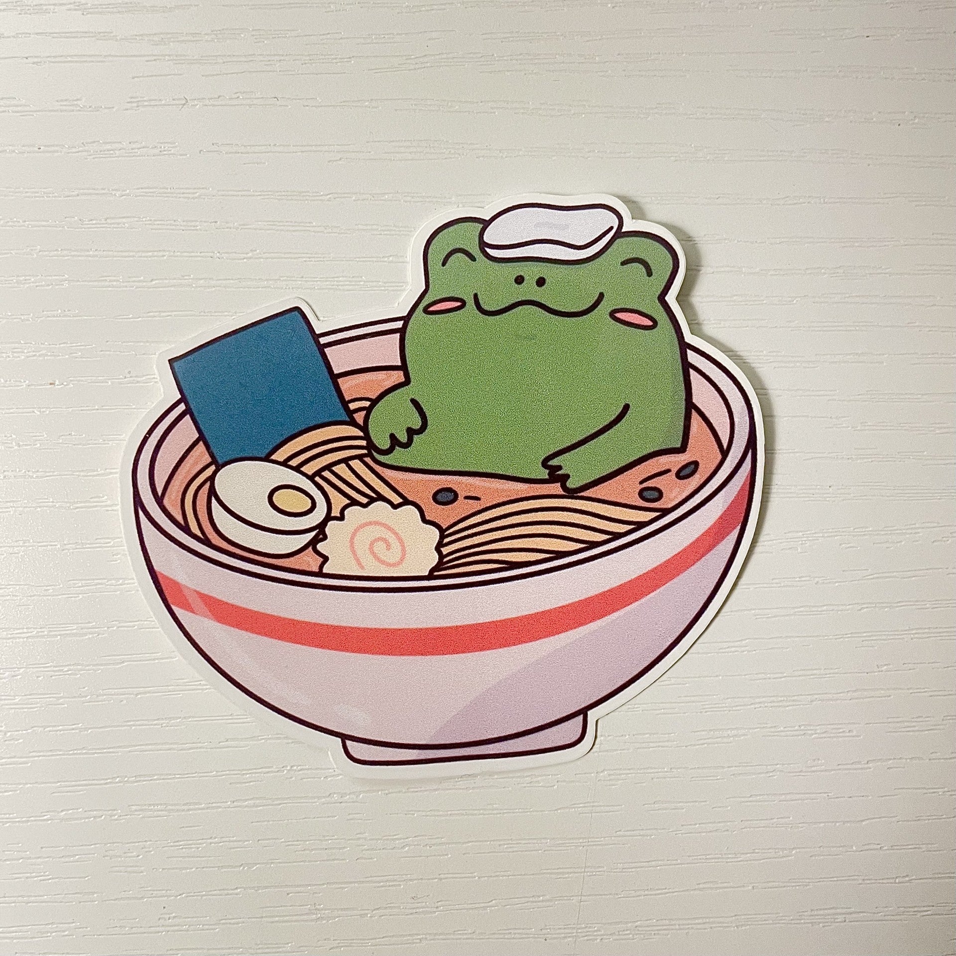 Cute green cartoon frog relaxing in a bowl of ramen noodles with a white towel on its head, a soft boiled egg, and fish cake.